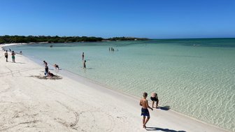 Ein sonniger Strand mit feinem, weißem Sand, wo Kinder spielen und im klaren Wasser planschen.