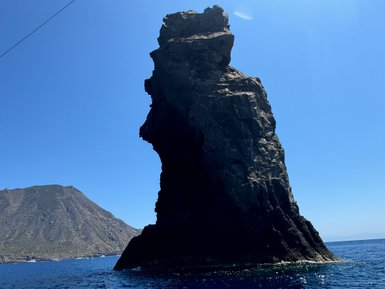 Ein markanter, dunkler Felsen erhebt sich majestätisch aus dem blauen Wasser, umgeben von sanften Hügeln im Hintergrund.