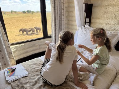 Zwei Mädchen sitzen auf einem Bett und betrachten Zeichnungen, während Elefanten durch das Fenster im Hintergrund sichtbar sind.
