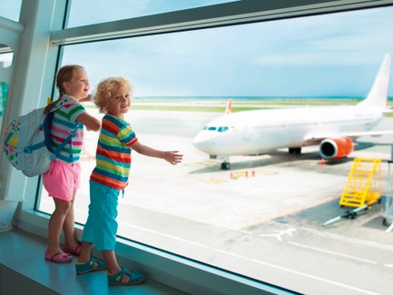 Zwei fröhliche Kinder stehen am Fenster eines Flughafens und beobachten ein Flugzeug auf dem Rollfeld.