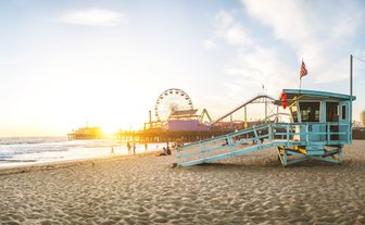 Ein Rettungsturm steht am Strand von Santa Monica, während die Sonne hinter dem Pier untergeht und die Wellen sanft an den Strand rollen.