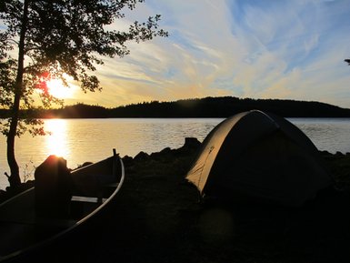 Ein Zelt steht am Ufer eines ruhigen Sees, während die Sonne hinter den Bäumen untergeht und den Himmel in warmen Farben färbt.