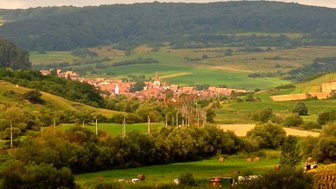 Eine malerische Landschaft mit einem kleinen Dorf, umgeben von sanften Hügeln und üppigem Grün unter einem bewölkten Himmel.
