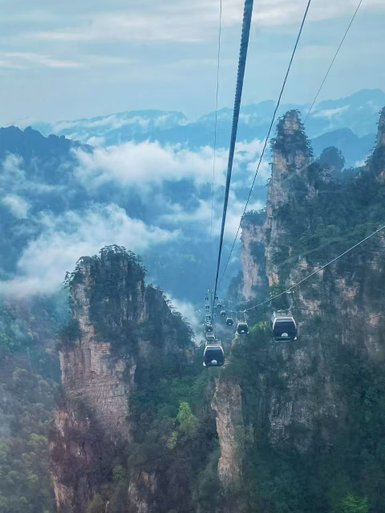 Eine Seilbahn schwebt über majestätische, bewaldete Berge, umgeben von mystischen Wolken und einem blauen Himmel.