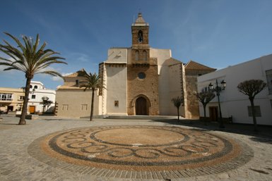 Eine historische Kirche mit einem markanten Glockenturm steht im Mittelpunkt eines gepflasterten Platzes, umgeben von Palmen.