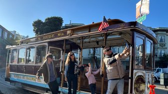 Eine Gruppe von vier Personen posiert fröhlich an einer historischen Kabelbahn in San Francisco, umgeben von sonnigem Wetter.