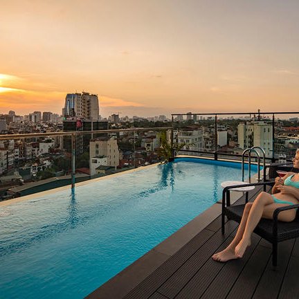 Eine Frau in einem blauen Badeanzug entspannt am Rand eines Infinity-Pools mit Blick auf die Stadt bei Sonnenuntergang.