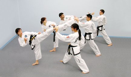 Sechs Taekwondo-Praktizierende in weißen Anzügen üben gemeinsam Techniken in einem hellblauen Raum.