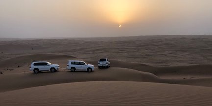 Drei weiße SUVs stehen auf sanften Sanddünen, während die Sonne am Horizont untergeht und den Himmel in sanften Farben färbt.
