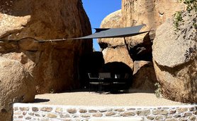 Eine gemütliche Sitzecke unter einem Sonnensegel, umgeben von beeindruckenden Felsen und klarem blauen Himmel.