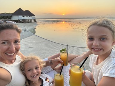Eine Frau und zwei Mädchen genießen fruchtige Cocktails am Strand, während die Sonne über dem Wasser untergeht.