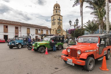 Bunte Jeeps parken auf einem Platz, umgeben von historischen Gebäuden und einer Kirche mit einer markanten Uhr.