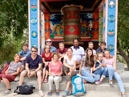 Eine Reisegruppe sitzt lächelnd vor einem bunten tibetischen Tempel mit einem großen Gebetsrad im Hintergrund.