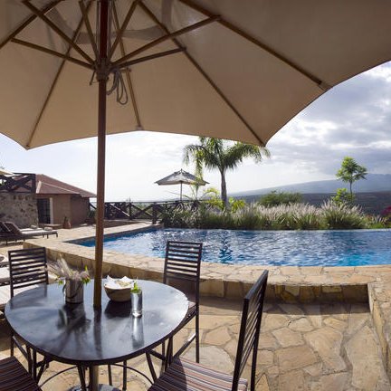 Eine einladende Poolterrasse mit Liegen, einem runden Tisch und Sonnenschirmen, umgeben von üppigem Grün und Bergen im Hintergrund.