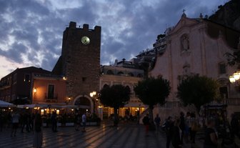 Der Platz in Taormina ist bei Dämmerung beleuchtet, mit einem historischen Turm und einer Kirche im Hintergrund.