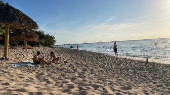 Zwei Kinder sitzen im Sand am Strand, während ein Junge im Hintergrund am Wasser steht und die Wellen beobachtet.