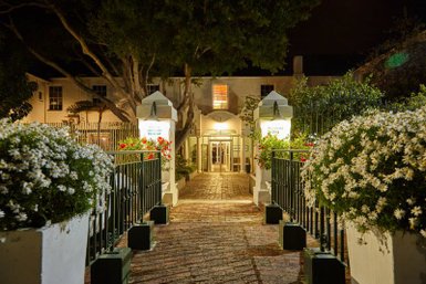 Der Eingangsbereich des Windsor Hotels in Hermanus ist nachts beleuchtet, umgeben von blühenden Blumen und grünen Bäumen.