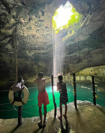 Zwei Jungen stehen am Rand eines Cenotes und schauen auf das glitzernde Wasser, während Lichtstrahlen von oben fallen.