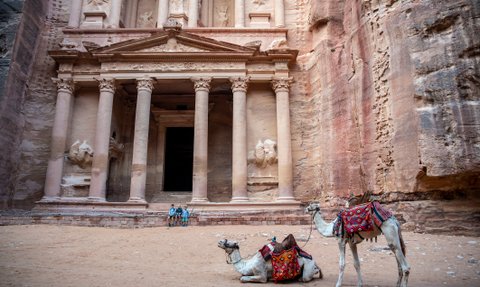 Zwei Kamele mit bunten Sätteln ruhen im Sand vor der beeindruckenden Fassade eines antiken Gebäudes in Petra.