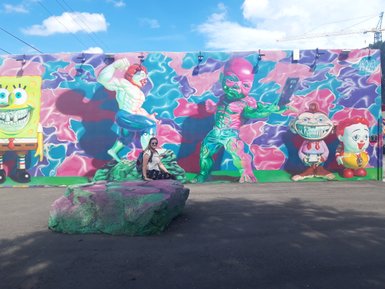 Eine Person sitzt auf einem bunten Felsen vor einer lebhaften Wandmalerei mit Cartoonfiguren in Wynwood, Miami.