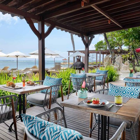 Ein einladendes Restaurant am Strand mit stilvollen Tischen und Stühlen, umgeben von Palmen und Sonnenschirmen.