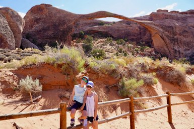 Ein Junge und ein Mädchen posieren lächelnd vor dem beeindruckenden Bogen im Arches Nationalpark, umgeben von rotem Sand.