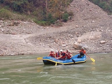 Eine Gruppe von sechs Personen paddelt in einem blauen Raft auf dem Trishuli-Fluss, umgeben von felsigen Ufern und Bäumen.