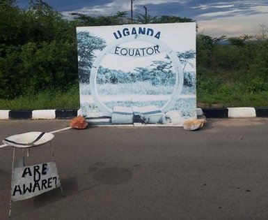 Ein großes, bemaltes Schild mit der Aufschrift 'UGANDA EQUATOR' steht vor einer grünen Landschaft unter einem bewölkten Himmel.