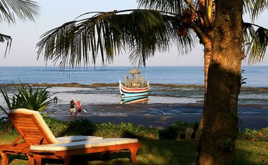 Ein ruhiger Strandblick mit einem traditionellen Boot im Wasser und einem Liegestuhl im Vordergrund, umgeben von Palmen.