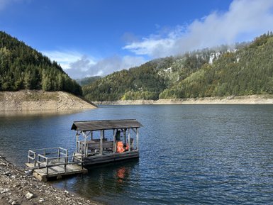 Ein Holzsteg ragt über das ruhige Wasser eines Stausees, umgeben von bewaldeten Hügeln und einem klaren blauen Himmel.