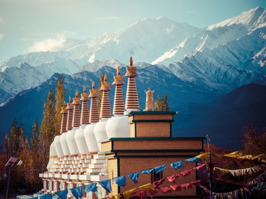 Eine Reihe von Stupas mit goldenen Spitzen steht vor majestätischen, schneebedeckten Bergen im Hintergrund.
