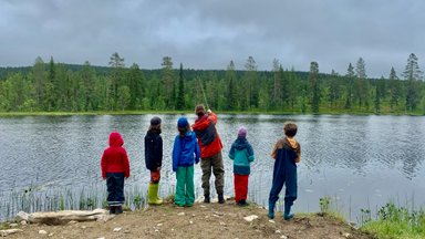 Sechs Kinder stehen am Ufer eines ruhigen Sees und beobachten einen Erwachsenen beim Angeln in der Natur.