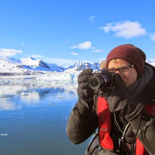 Eine Person mit einer Kamera steht am Wasser und fotografiert die beeindruckende Berglandschaft im Hintergrund.