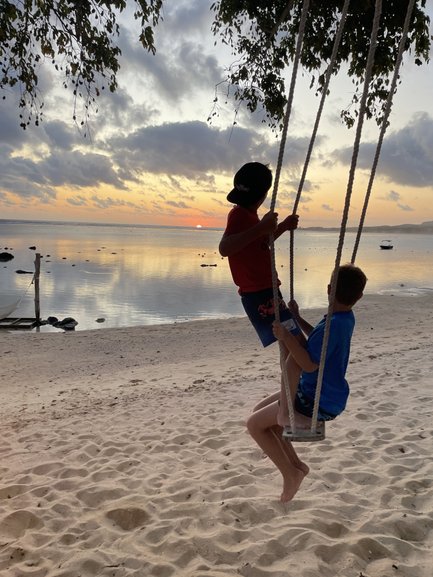 Zwei Kinder schwingen fröhlich auf einer Schaukel am Strand, während die Sonne hinter dem Horizont untergeht.