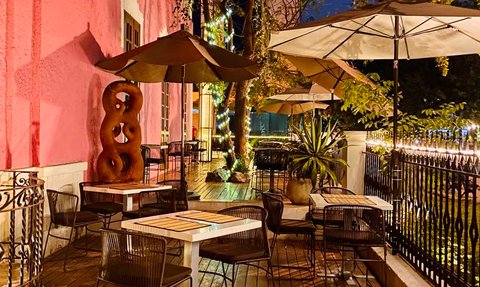 Eine einladende Terrasse mit Tischen und Stühlen, umgeben von einem pinken Gebäude und sanfter Beleuchtung in der Dämmerung.