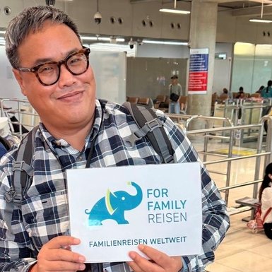Ein lächelnder Reiseleiter hält ein Schild mit der Aufschrift 'FOR FAMILY REISEN' am Flughafen in einer belebten Umgebung.