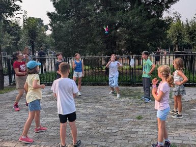 In einem Park spielen mehrere Kinder aktiv miteinander, während ein Erwachsener lächelnd zuschaut und die Atmosphäre genießt.