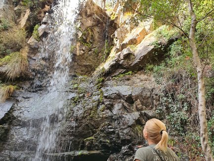 Eine Frau steht vor einem Wasserfall, der über eine felsige Klippe fließt, umgeben von üppigem Grün und Bäumen.