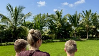 Eine Frau und zwei Kinder stehen vor einem üppigen Reisfeld, umgeben von Palmen und einem strahlend blauen Himmel.