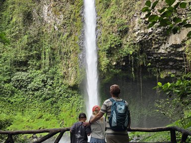 Eine Familie steht am Rand einer Aussichtsplattform und bestaunt einen majestätischen Wasserfall, umgeben von üppigem Grün.