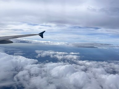 Der Blick aus dem Flugzeug zeigt die Wolkenlandschaft unterhalb, mit einem Flugzeugflügel im Vordergrund.