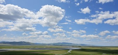 Eine weite, grüne Landschaft unter einem strahlend blauen Himmel mit vielen weißen Wolken und sanften Hügeln im Hintergrund.
