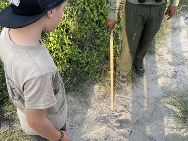 Ein Junge mit einer Kappe beobachtet aufmerksam die Fußabdrücke im Sand, während ein Erwachsener mit einem Stock daneben steht.