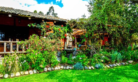 Ein üppiger Garten umgibt eine charmante Hacienda, mit bunten Blumen und einer Statue im Mittelpunkt der Szene.