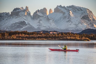 Ein Kajakfahrer paddelt ruhig auf einem glatten See, umgeben von majestätischen, schneebedeckten Bergen im Hintergrund.