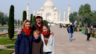 Eine Familie posiert fröhlich vor dem Taj Mahal, umgeben von grünen Bäumen und einem klaren Himmel.