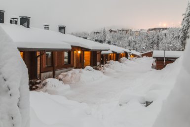 Eine verschneite Landschaft mit Holzchalets, die sanft im Winterlicht leuchten, umgeben von schneebedeckten Bäumen.