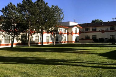 Ein helles Hotelgebäude mit orangefarbenen Akzenten, umgeben von gepflegtem Grün und hohen Bäumen im Vordergrund.