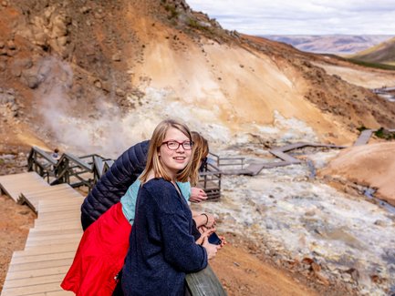 Eine junge Frau mit Brille lächelt, während sie an einem Geländer in einem geothermalen Gebiet steht, umgeben von dampfenden Quellen.
