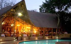 Ein stilvolles Safari-Lodge mit einem großen Strohdach, beleuchtet von warmem Licht, umgeben von Bäumen und einem Pool.